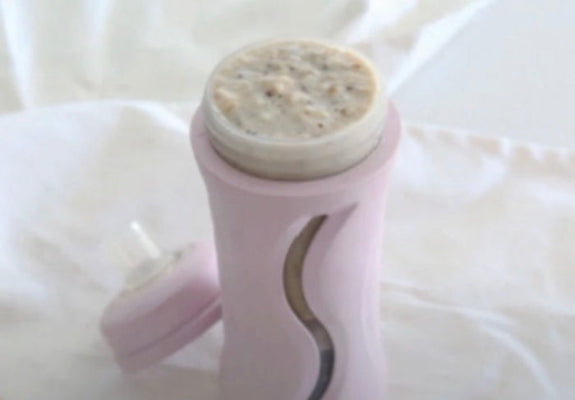 A pink baby bottle with a clear cap removed and lying beside it, filled with a beige textured food or powder, set against a soft white fabric background.