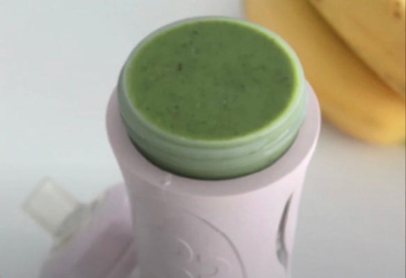 A close-up of a light pink portable blender cup filled with a green smoothie, with a banana blurred in the background on a white surface.