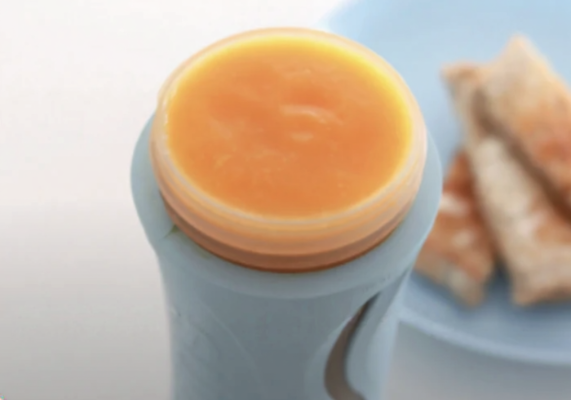 A close-up of a light blue silicone baby bottle with a clear lid filled with orange puree, with a plate of crackers blurred in the background.